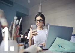 Une femme assise à un bureau consulte son téléphone devant un ordinateur portable