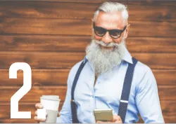 Un homme âgé à la barbe blanche, lunettes de soleil et bretelles, tient un café et un téléphone