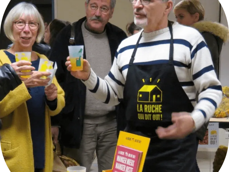 Un groupe de personnes souriantes trinquent lors d'une distribution de produits locaux, tabliers jaunes