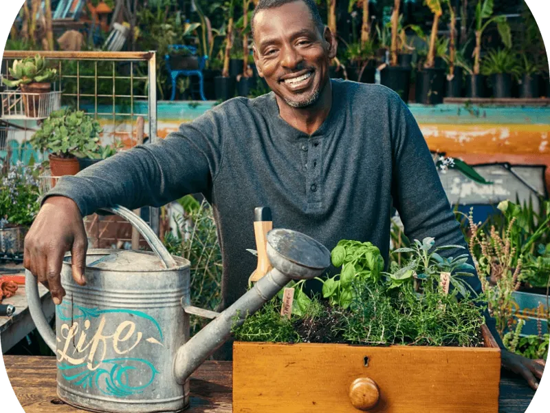 Un homme souriant arrose un potager surélevé en bois dans un jardin verdoyant