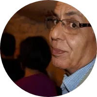 Portrait de Brahim, cheveux gris et lunettes, chemise à carreaux, dans une pièce sombre