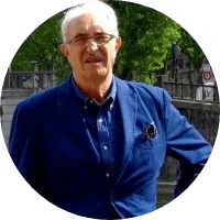 Portrait d'Alain, cheveux blancs, lunettes et veste bleue, debout dans une rue arborée