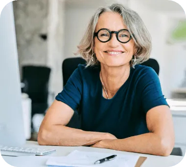 Une femme aux cheveux gris, souriante, assise à son bureau devant un ordinateur portable